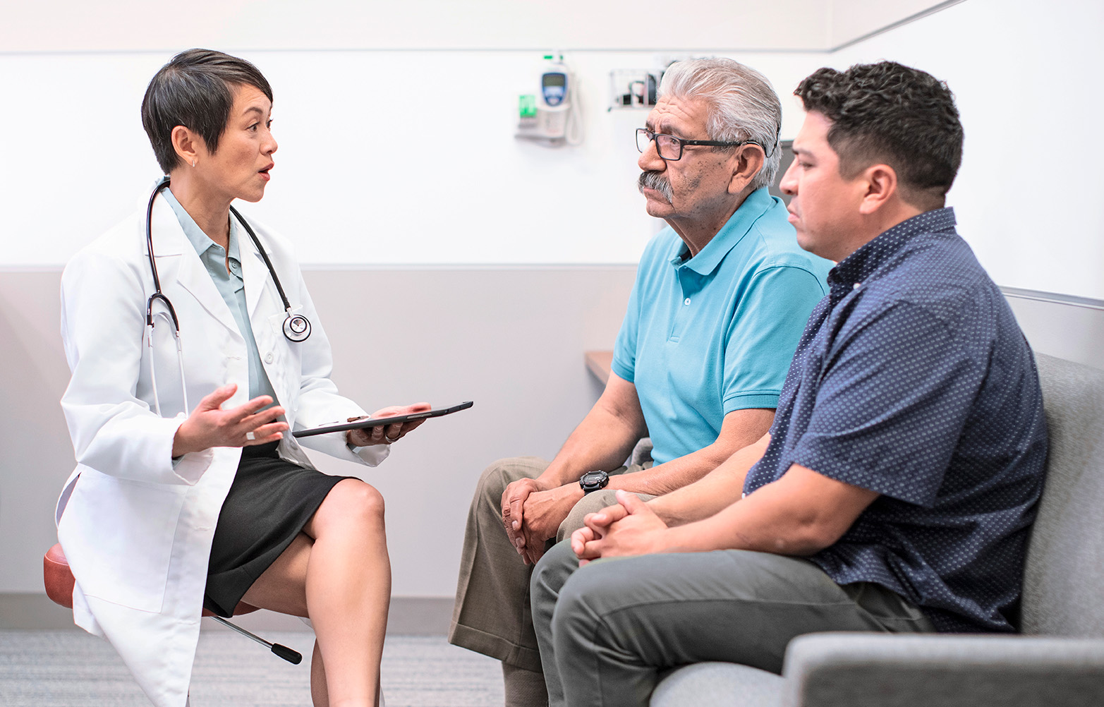 Female-presenting Asian doctor sits across from two male-presenting patients, a middle-aged person and an older person.
