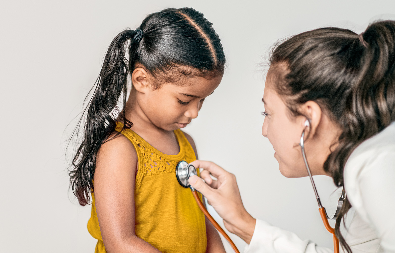 Female-presenting doctor leaning in with a stethoscope to hear a female-presenting child's heartbeat.