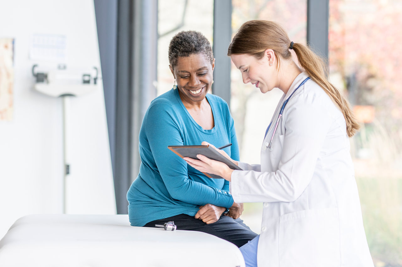 A senior patient of African decent, sits up on an exam table across from her female doctor as they discuss her medical concerns.  She is dressed casually and listening attentively as the doctor talks with her.  Her female doctor is wearing a white lab coat and has a tablet in her hands as she takes notes throughout.